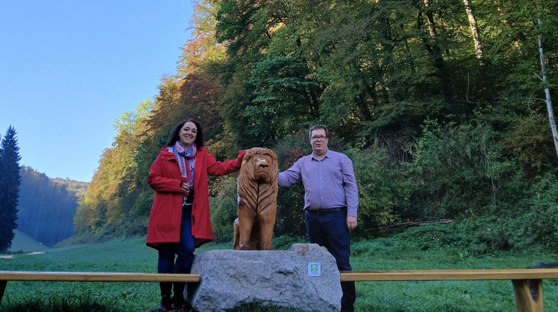 Annette Kölle und Tourismusmangager Florian Pointke stehen neben der neuen Löwenskulptur aus Holz am Filsursprung