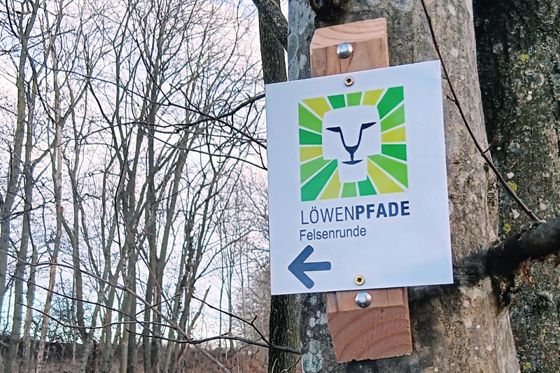Wanderzeichenschild im Wald mit einem Löwenkopf-Symbol.